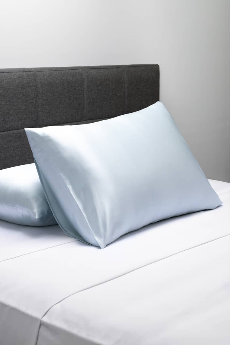 Smooth silver silk pillowcases on a bed with dark gray upholstered headboard.