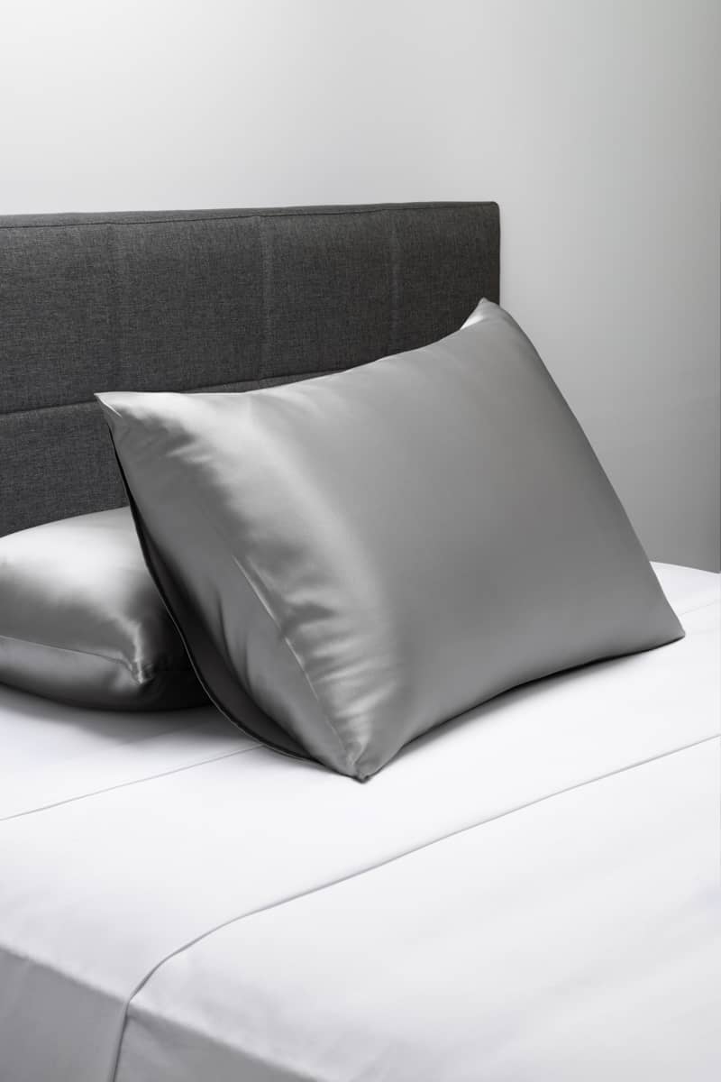 Silver silk pillowcases with a smooth, glossy finish on a gray upholstered bed.